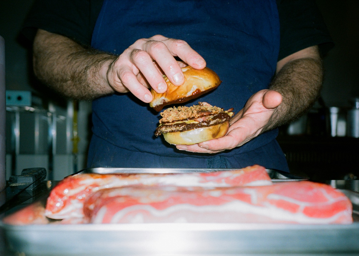 Un cuisinier referme un burger au bacon derrière un plateau de poitrine crue, Howard Original