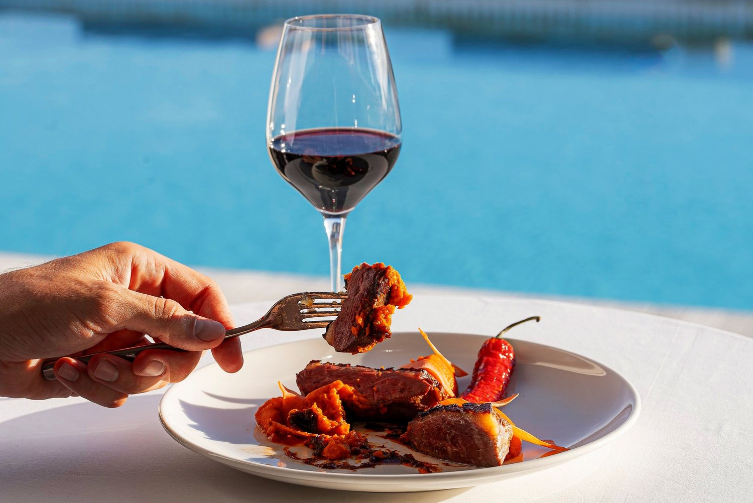 Une fourchette soulève une bouchée de viande aux poivrons, verre de rouge face à la piscine, Nhow