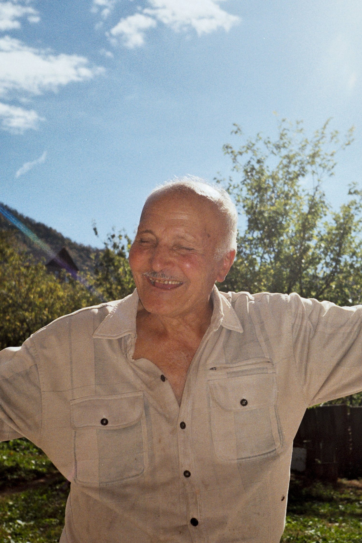 Portrait d'un homme âgé riant dans un jardin, mains sur les hanches, montagne au fond