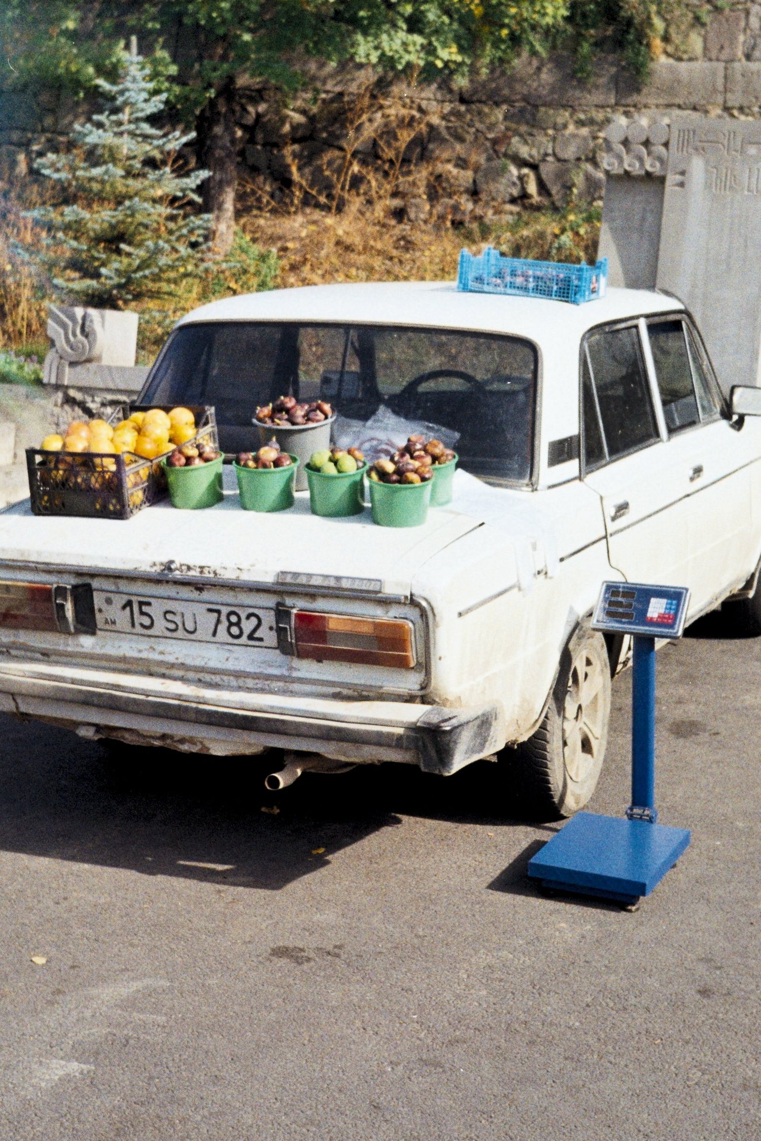 Vente de fruits sur le coffre d'une vieille berline blanche, balance posée au bord de la route