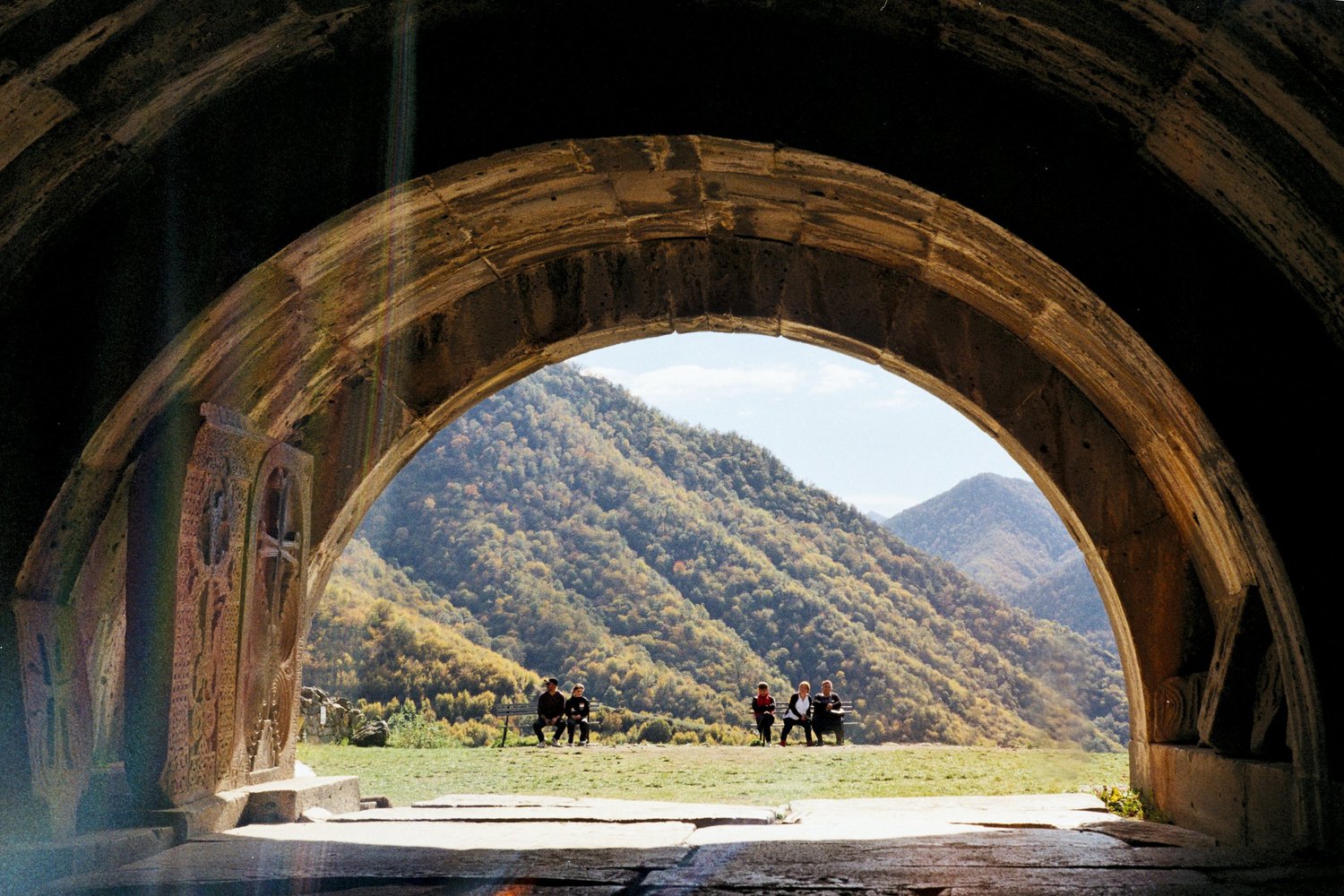 Vallée boisée encadrée par la grande arche de pierre d'un monastère, visiteurs assis dehors