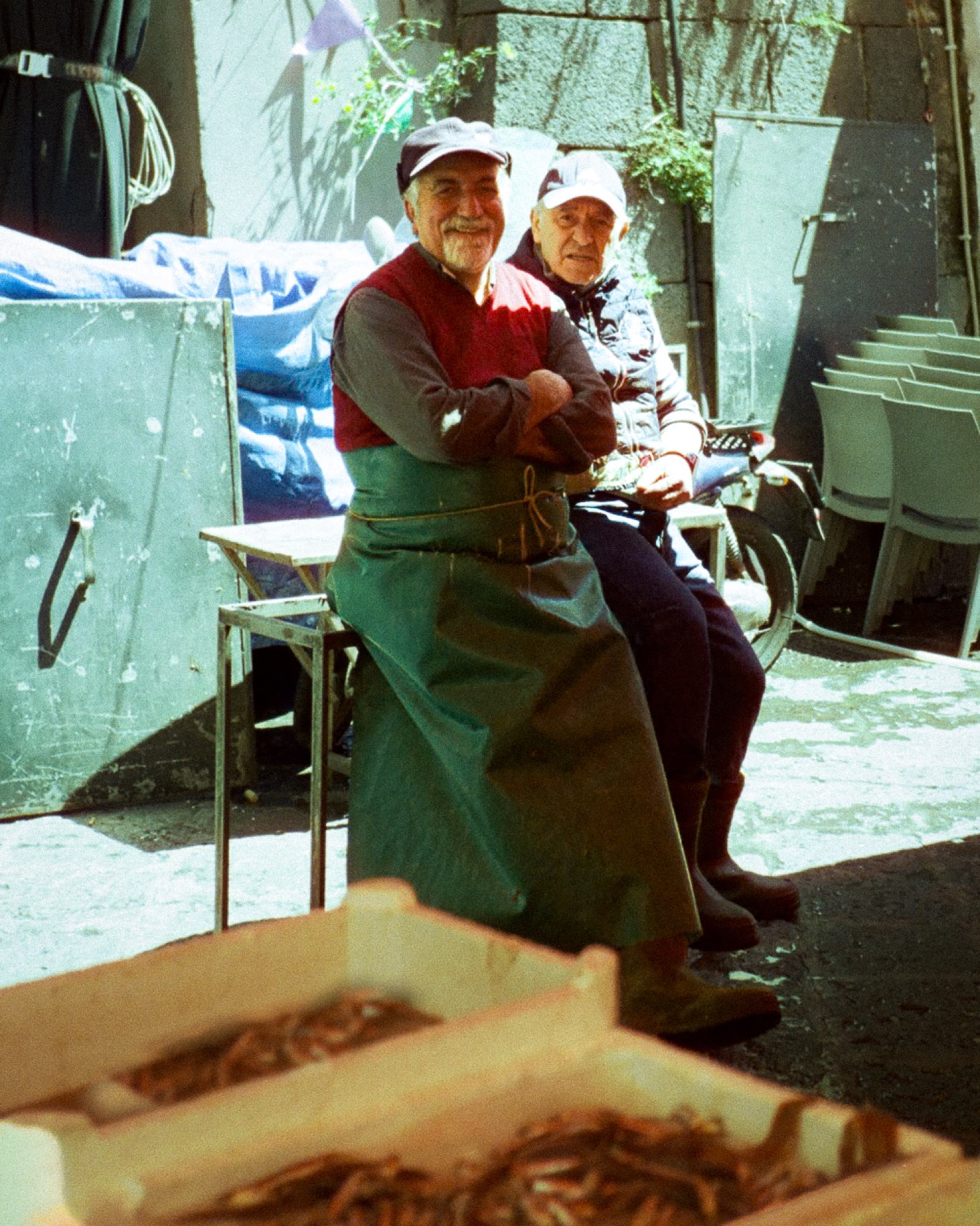 Deux poissonniers en tablier vert assis au soleil devant leurs cageots, marché sicilien